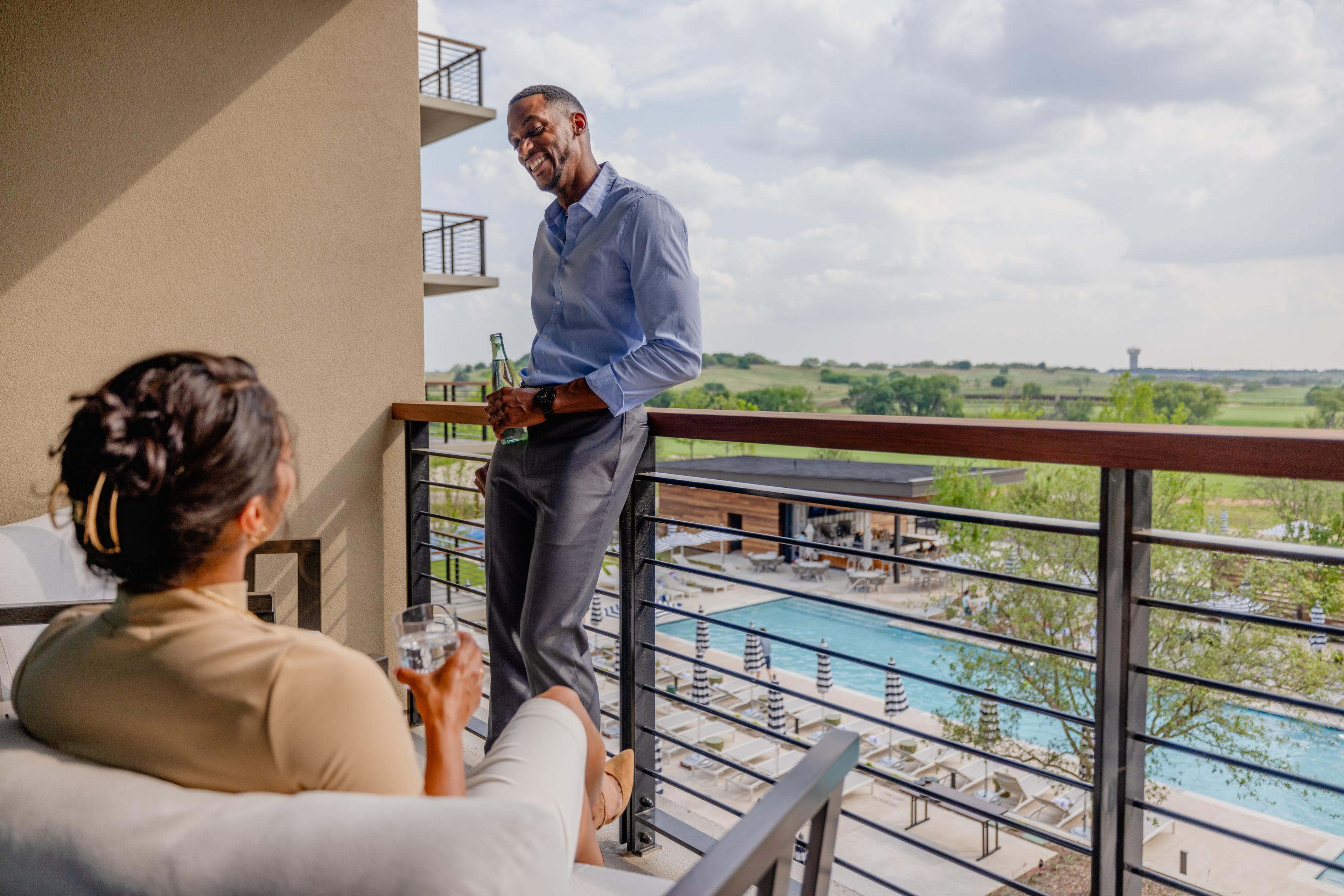 A man leaning against a balcony railing having a conversation with a seated woman as they both enjoy a drink.