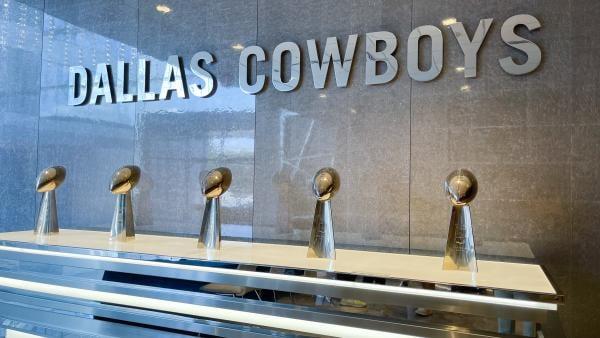 A wall with Dallas Cowboys written on it and 5 super bowl trophies on a ledge